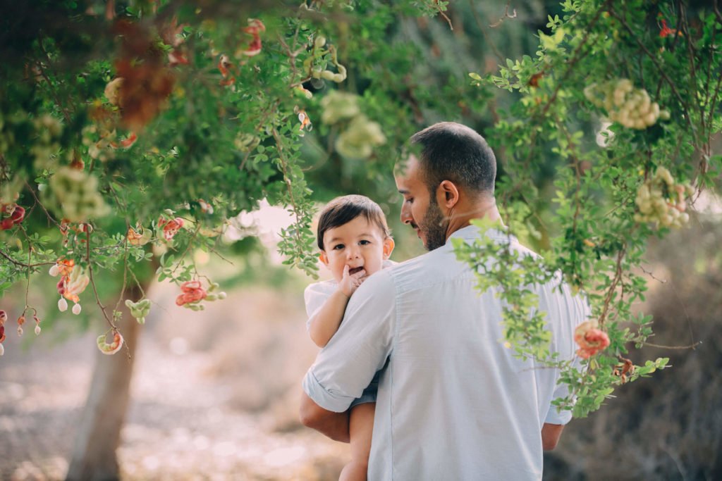 A dad holds his baby boy. They are surrounded by beautiful trees with orange flowers in Abu Dhabi