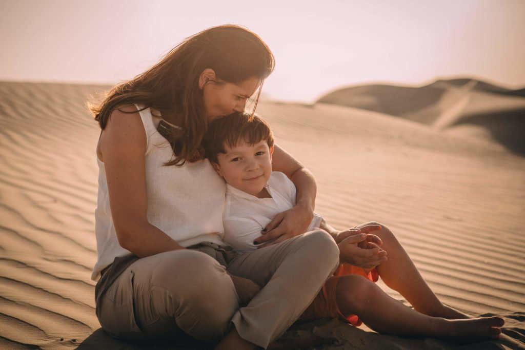 A mom kisses her son. They are in the desert dunes of Dubai
