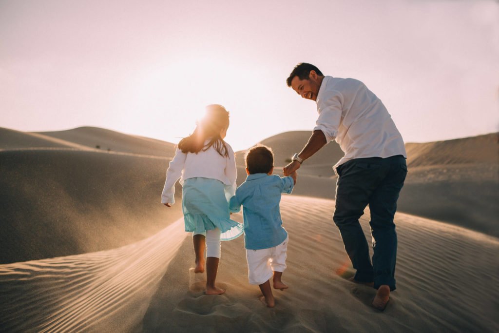 A dad holds the hands of his little boy and girl climbing the sand dunes during sunset