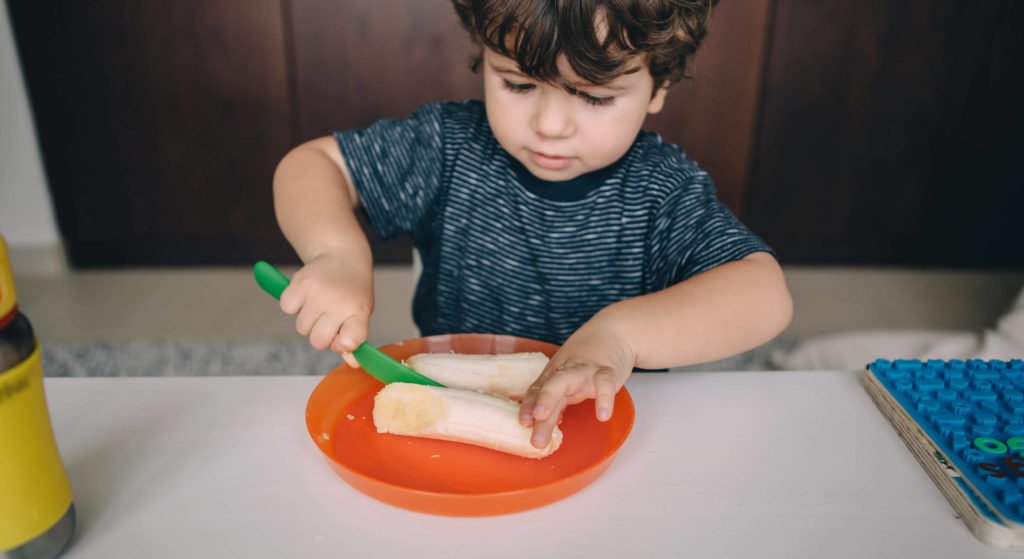 A toddler cuts his banana with a green plastic knife
