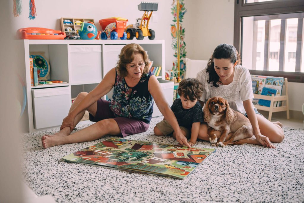 Grandmother shows a big book to her grandson while the mum looks at them with her dog in her lap